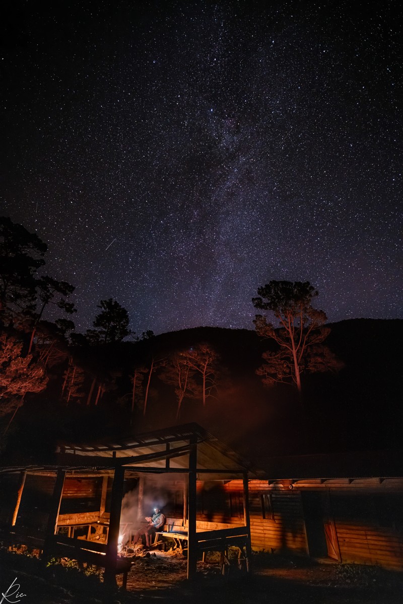 A small wooden structure glows beneath a star-filled sky.