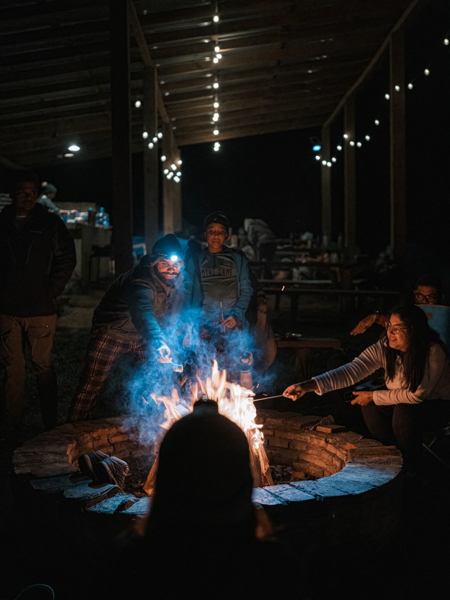 People gather around a fire under a roof strung with lights.
