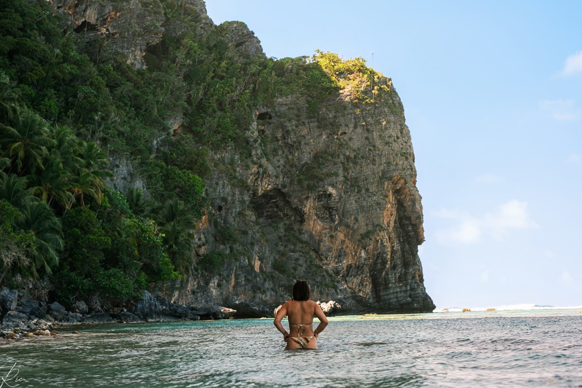 A figure sits in shallow coastal water below a tall rock face.