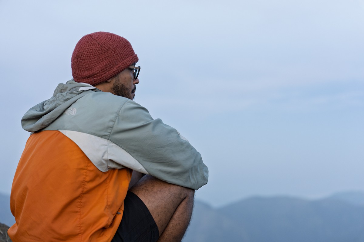 A seated figure in an orange layer looks across distant mountains.