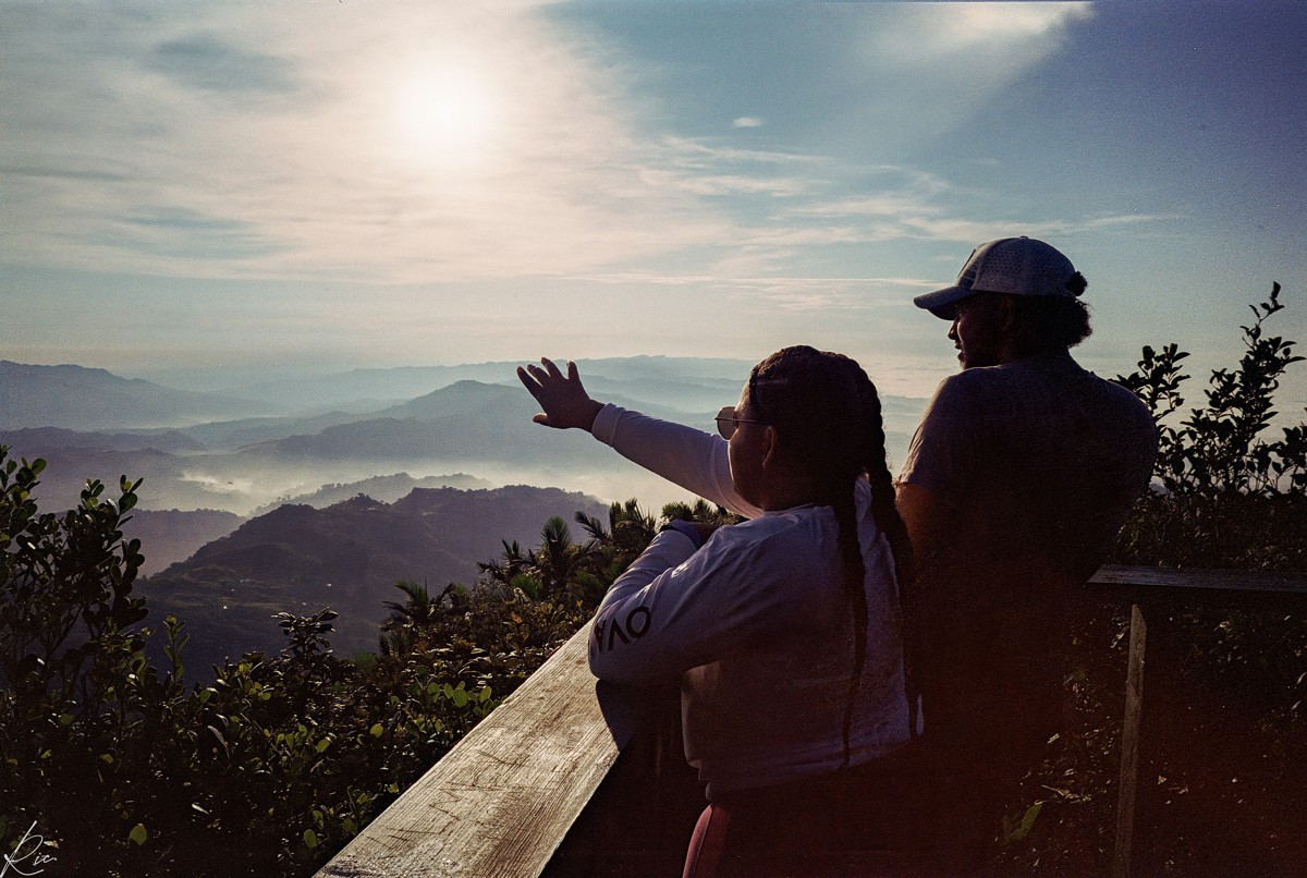 Two figures stand at a lookout while one gestures toward the mountains.