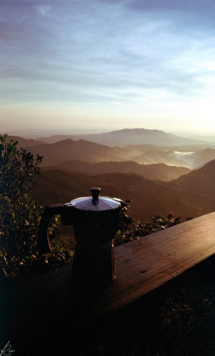 A dark cup rests on a railing against sunlit mountain layers.