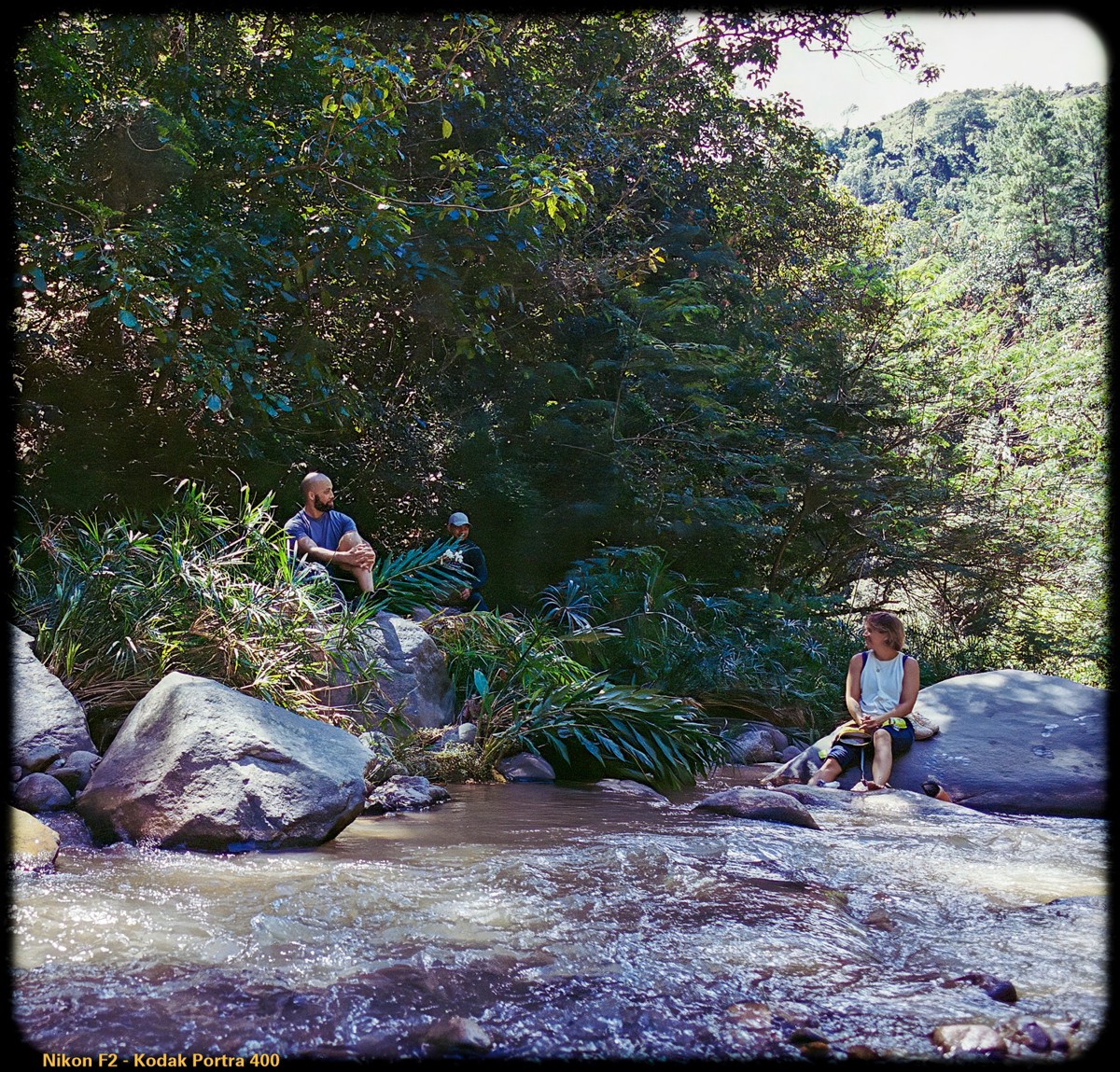 People pause among boulders beside a shaded stream.