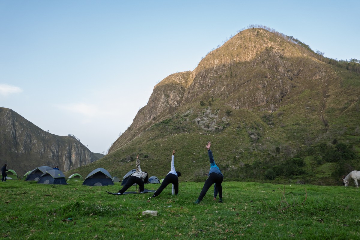 Campers stretch on grass below a steep mountain face.