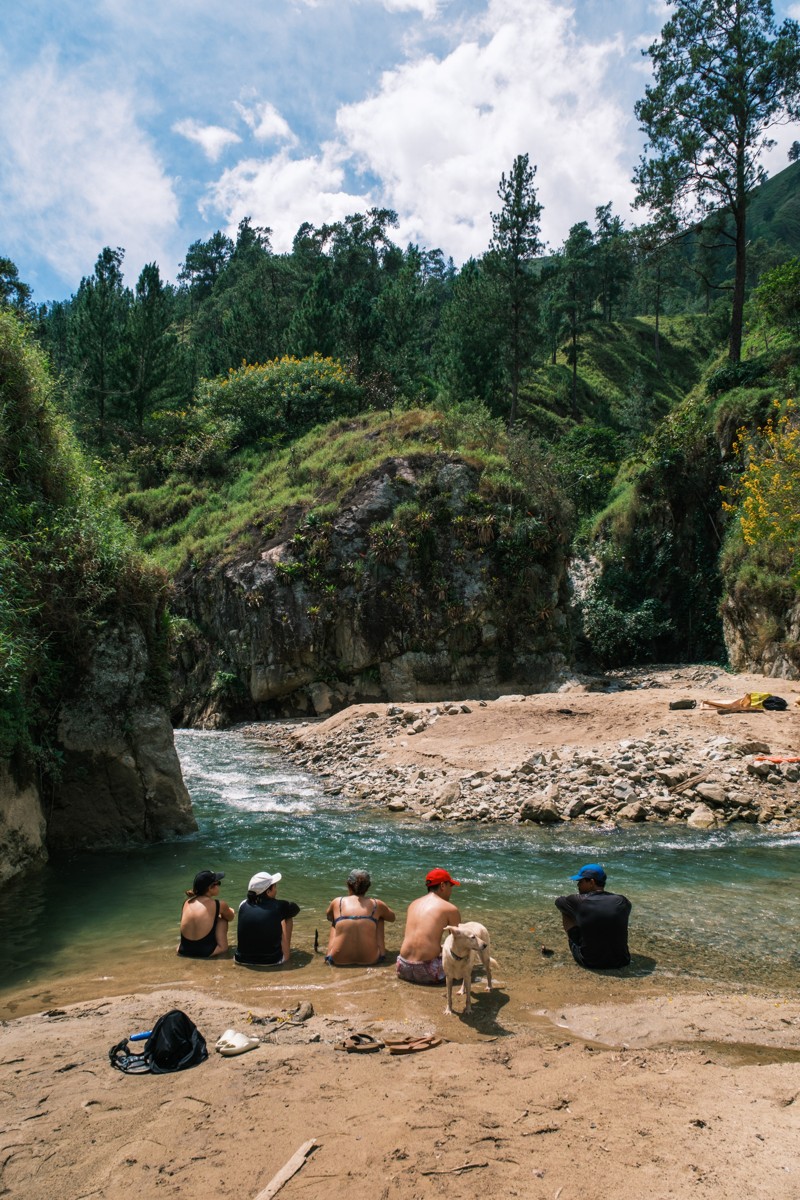 A group gathers at the edge of a clear pool below rocky banks.