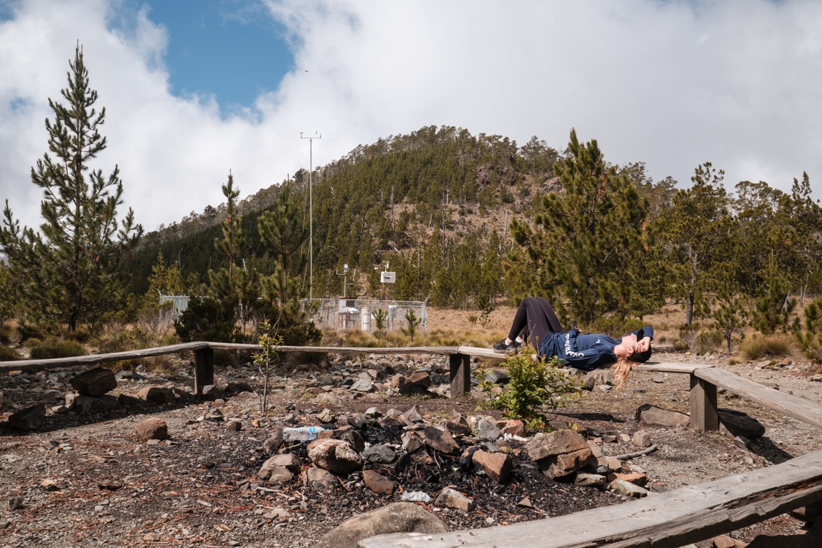 A person reclines beside a stone fire circle and timber benches.