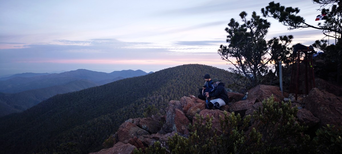 A figure sits on rocks overlooking layered mountains at dusk.