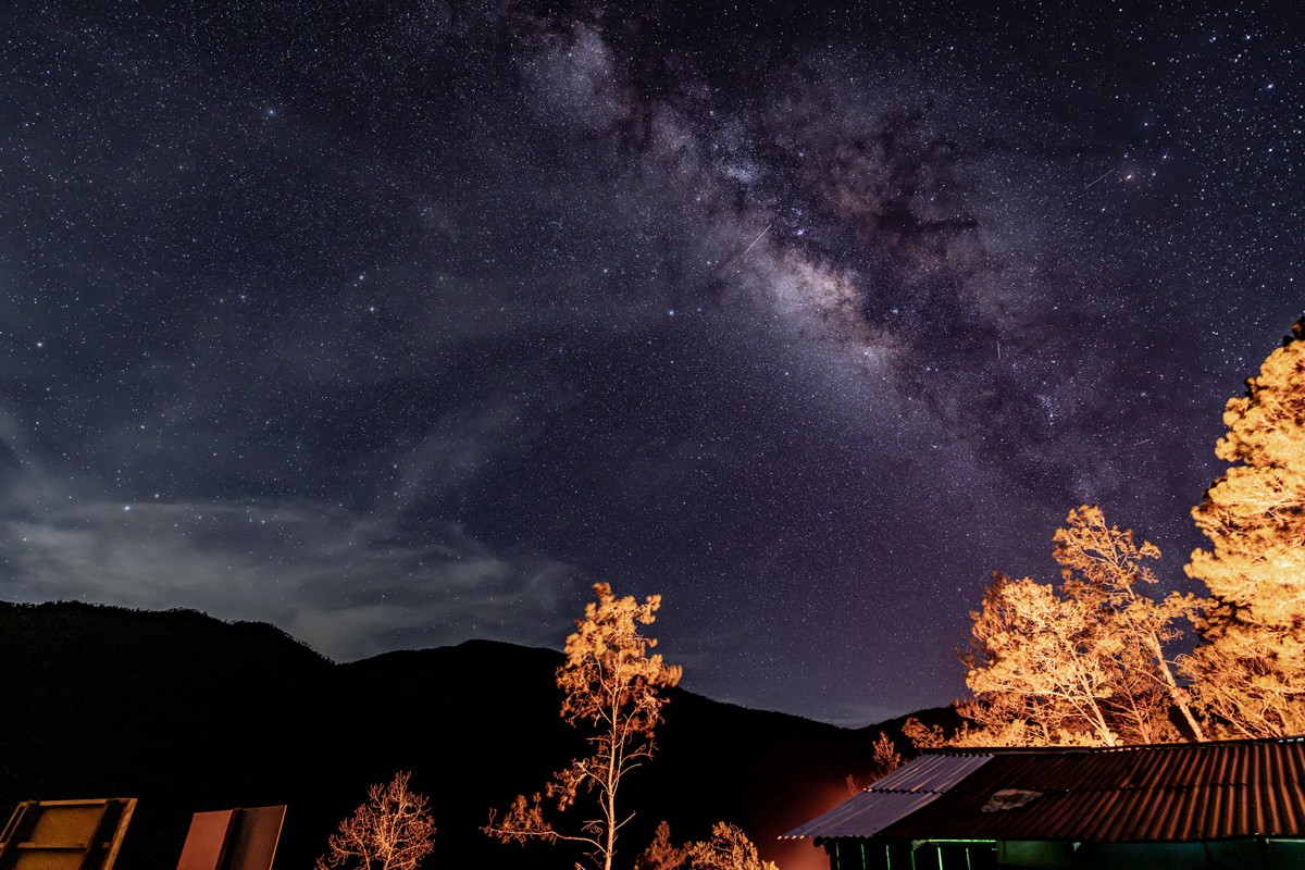 The Milky Way rises above a dark mountain silhouette and lit trees.