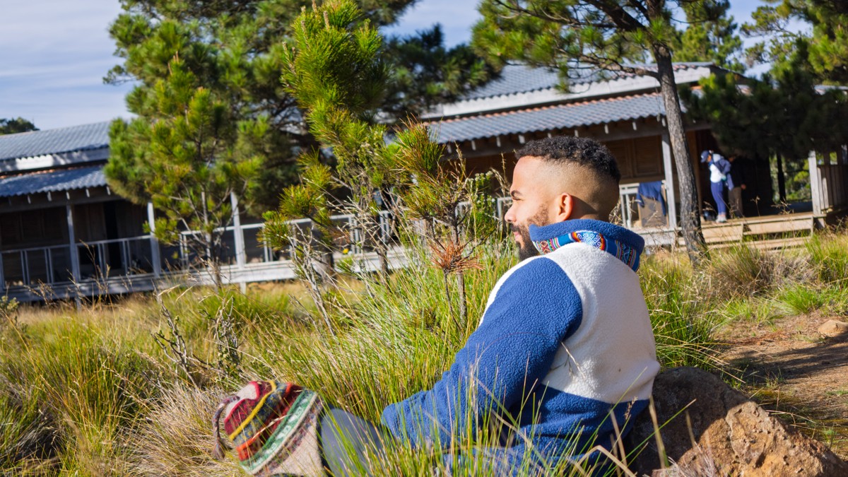 A man sits in the grass beside a timber shelter.