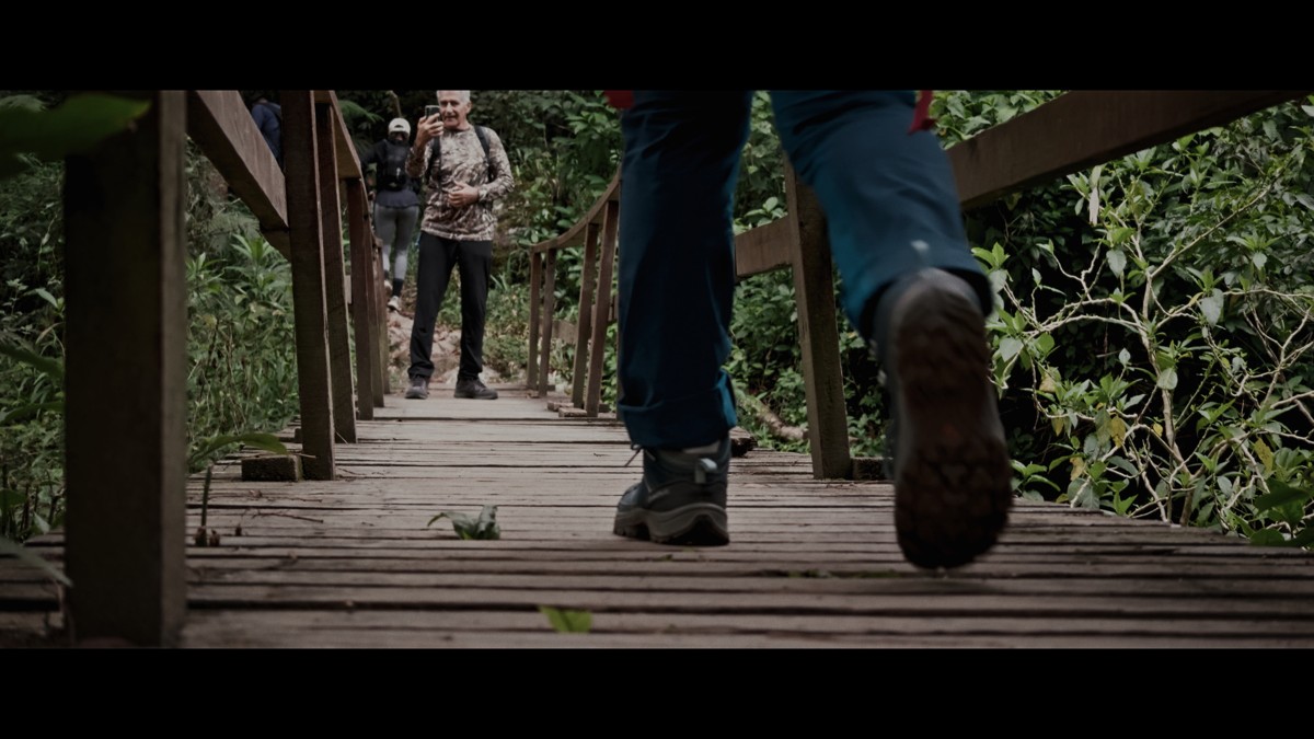 A low camera angle follows boots across a wooden bridge.