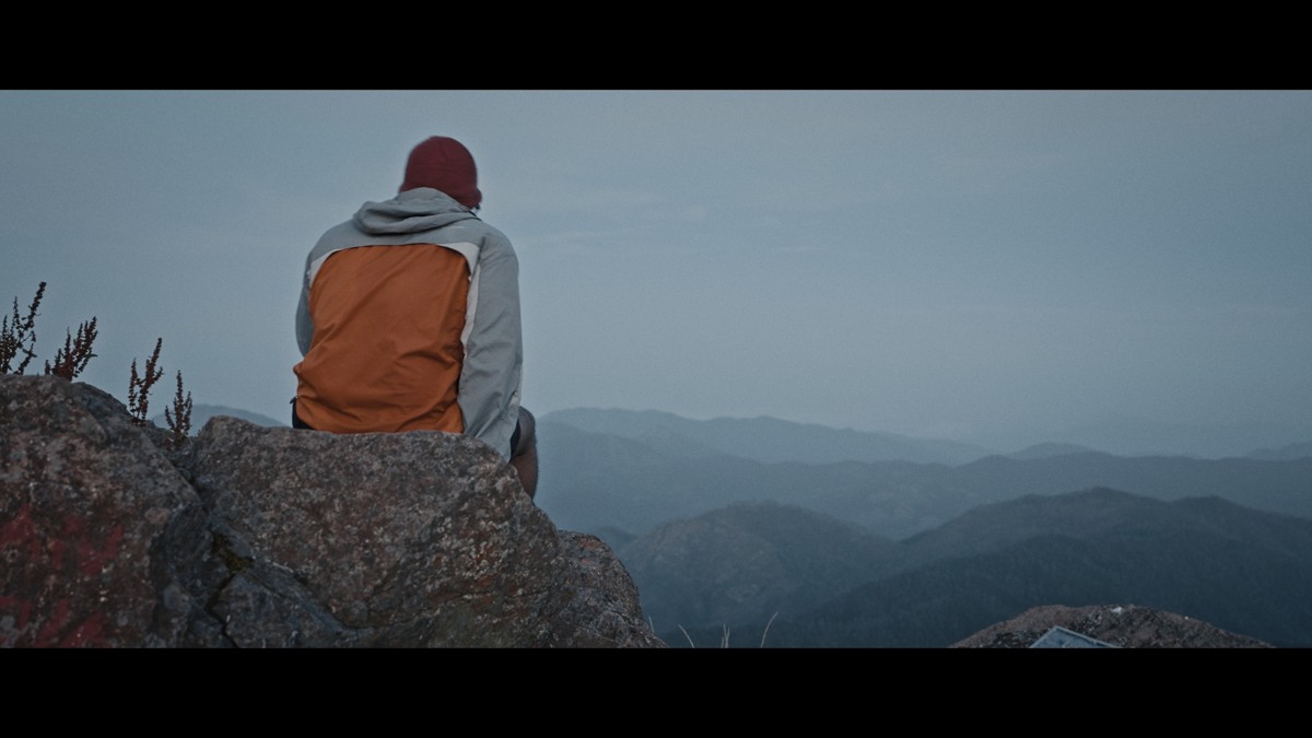 A person in an orange layer sits on a rock above distant ridges.