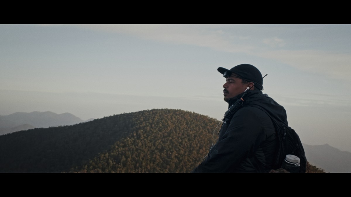 A hiker in a dark cap rests with the landscape behind him.