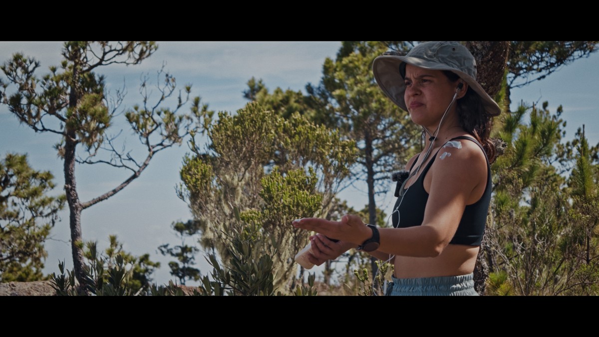 A hiker gestures beside sunlit pine branches.