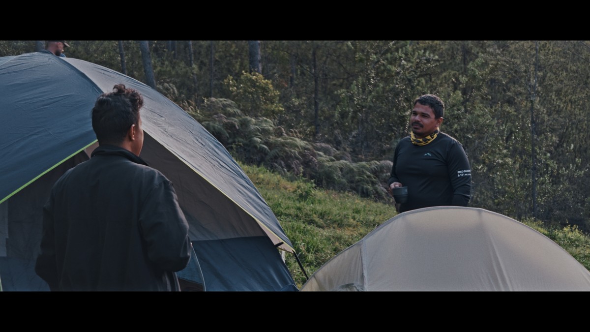 Two hikers stand between pitched tents at the forest edge.