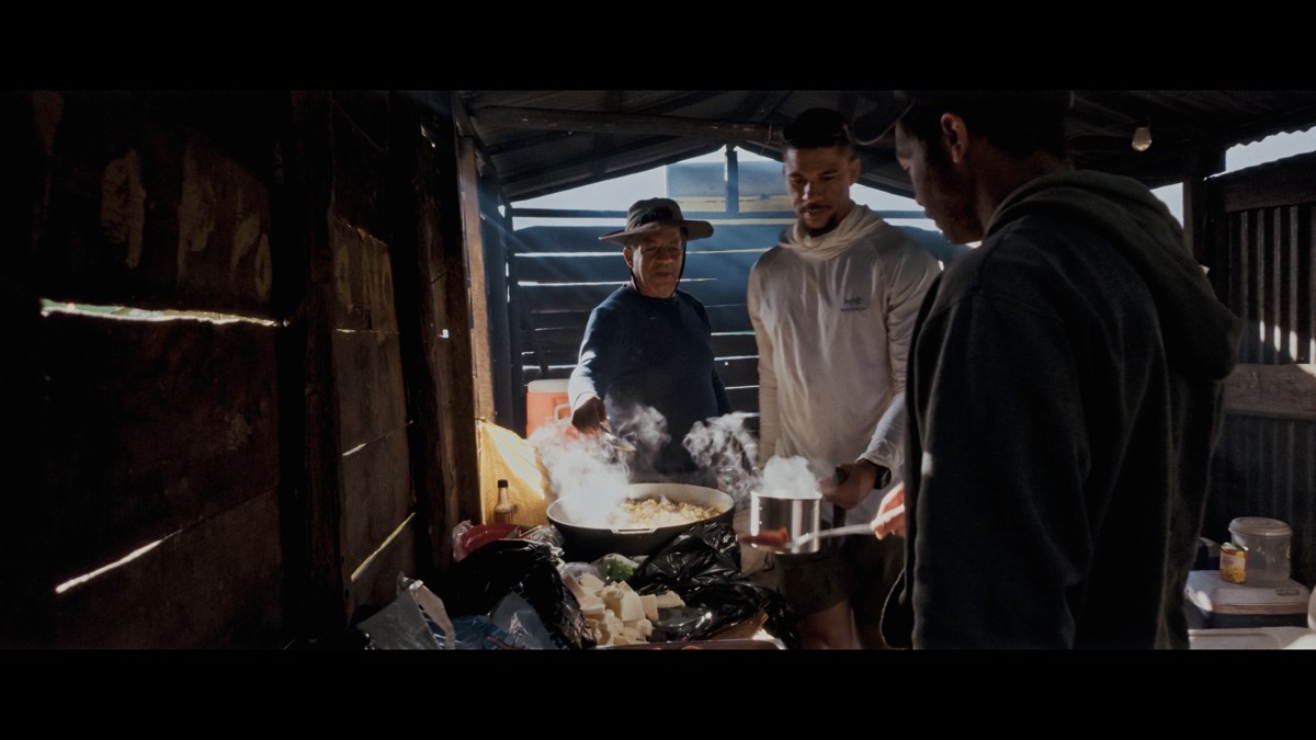People gather around a steaming pot inside a timber shelter.