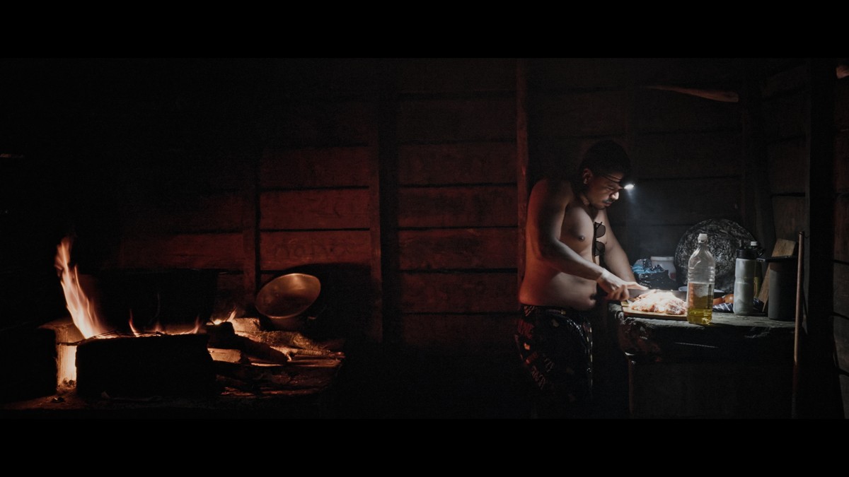 A person prepares food beside a small fire in a dark wooden room.