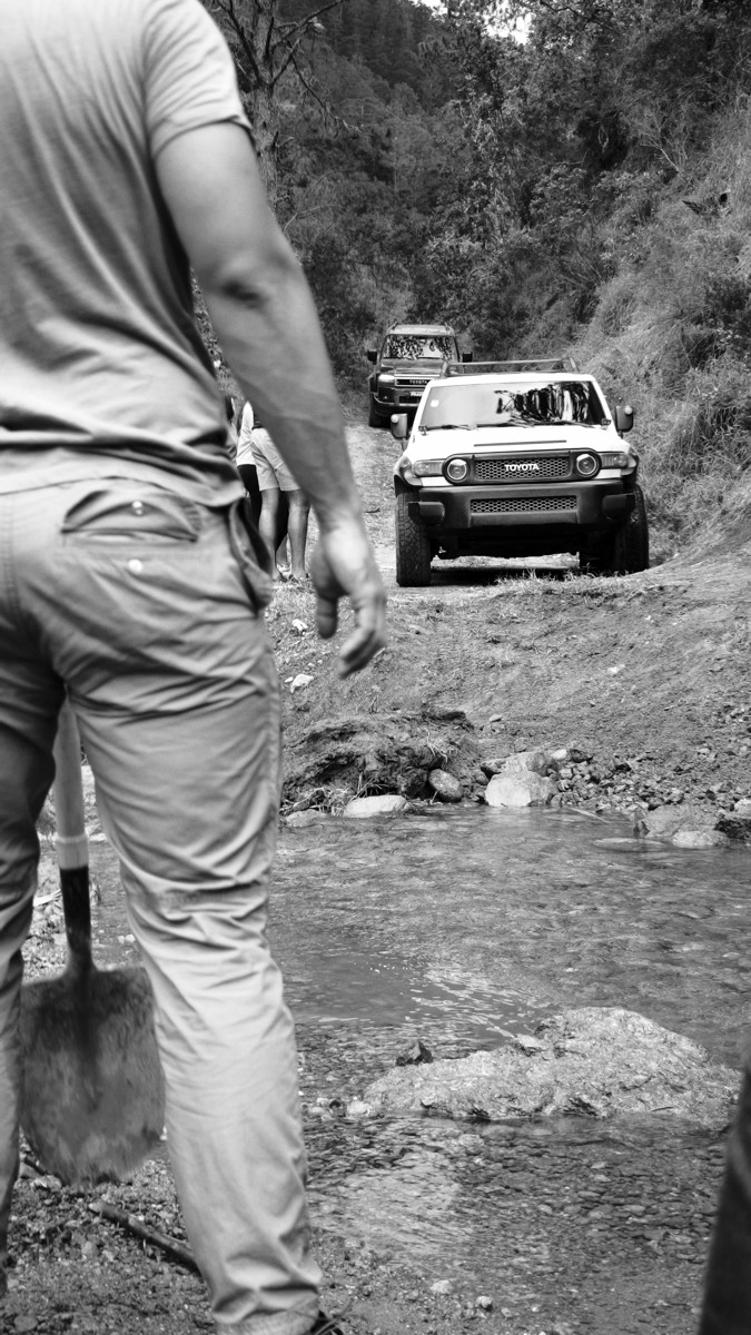 A standing figure watches a vehicle on a rocky track in monochrome.