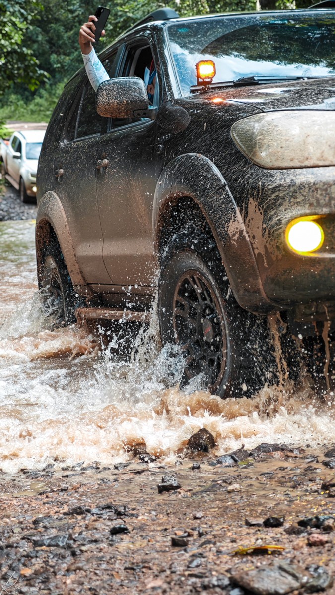A dark vehicle pushes through a shallow stream with its lights on.