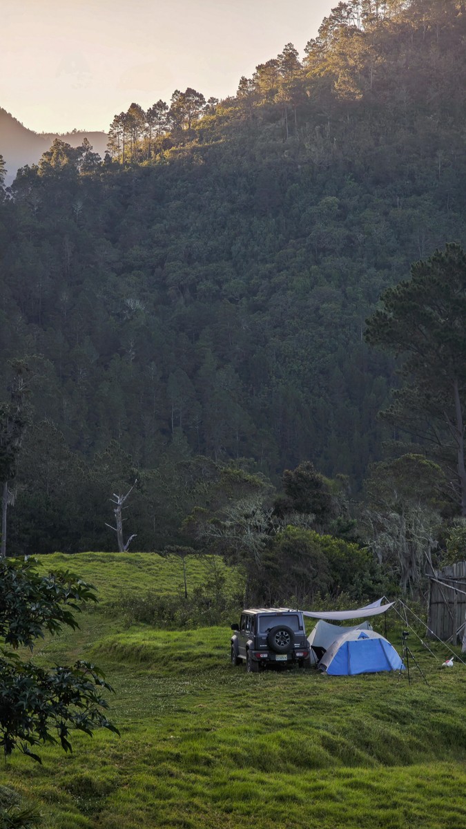 A tent and vehicle occupy a clearing beneath a high wooded slope.